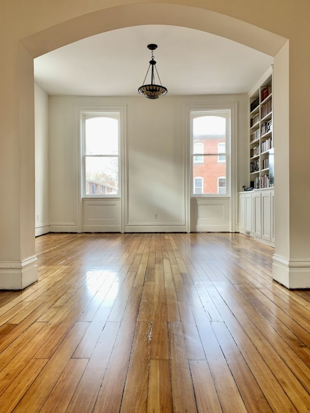 An arched entryway leads to a sunlit room with hardwood floors, two large windows, a ceiling light, and built-in shelves.