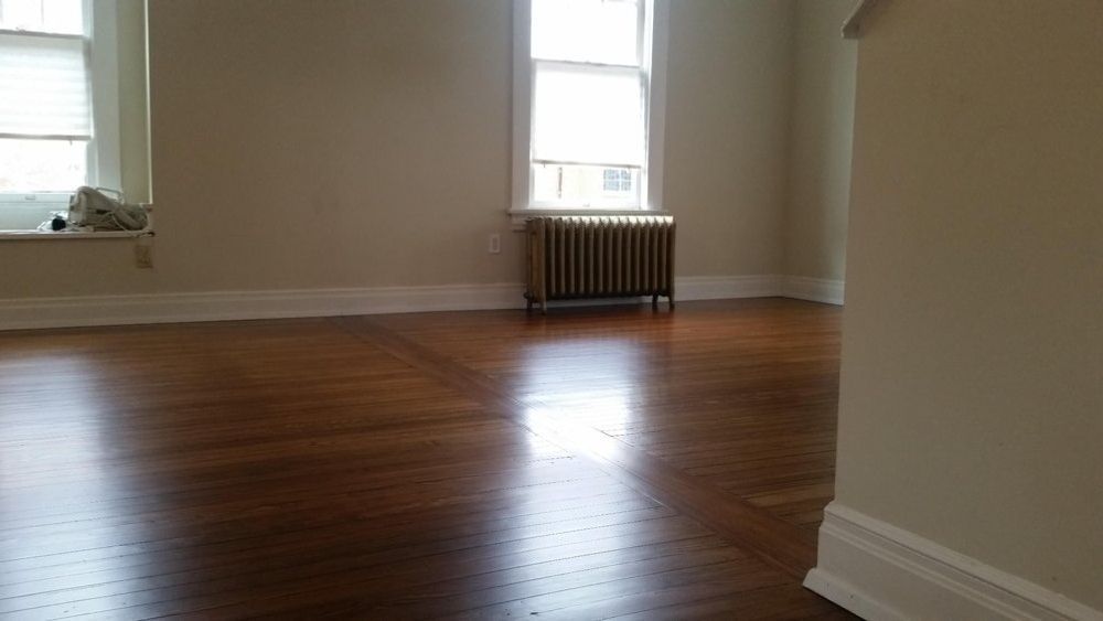An empty room with polished hardwood floors, cream-colored walls, two windows, and a vintage metal radiator.