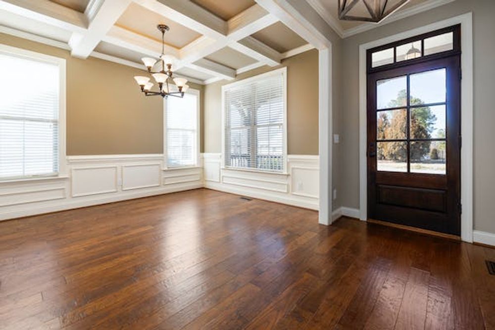 A brightly lit dining area with dark wood floors, coffered ceilings, wainscoting, and a glass-paneled dark wood front door.