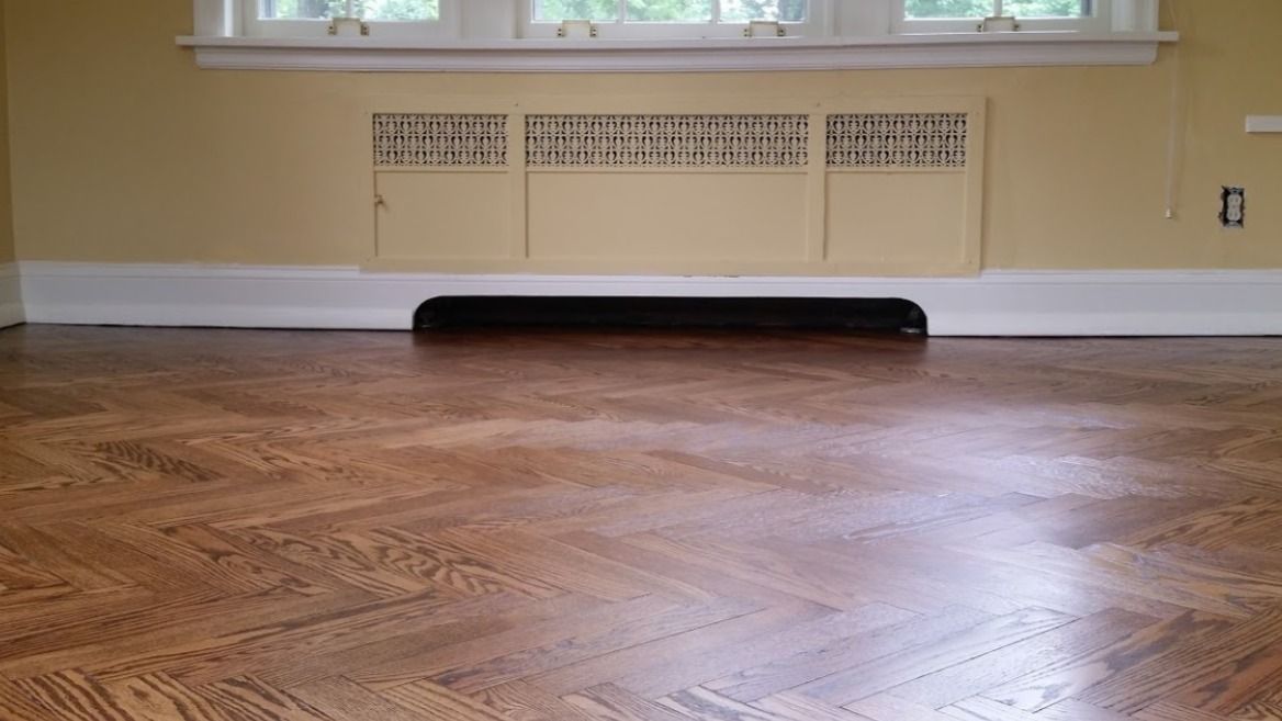 A room with a polished, patterned wood floor and a beige radiator cover centered below a window.