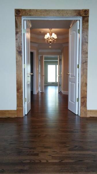A doorway with thick rustic wooden trim leads into a long hallway with dark hardwood floors and a hanging chandelier.