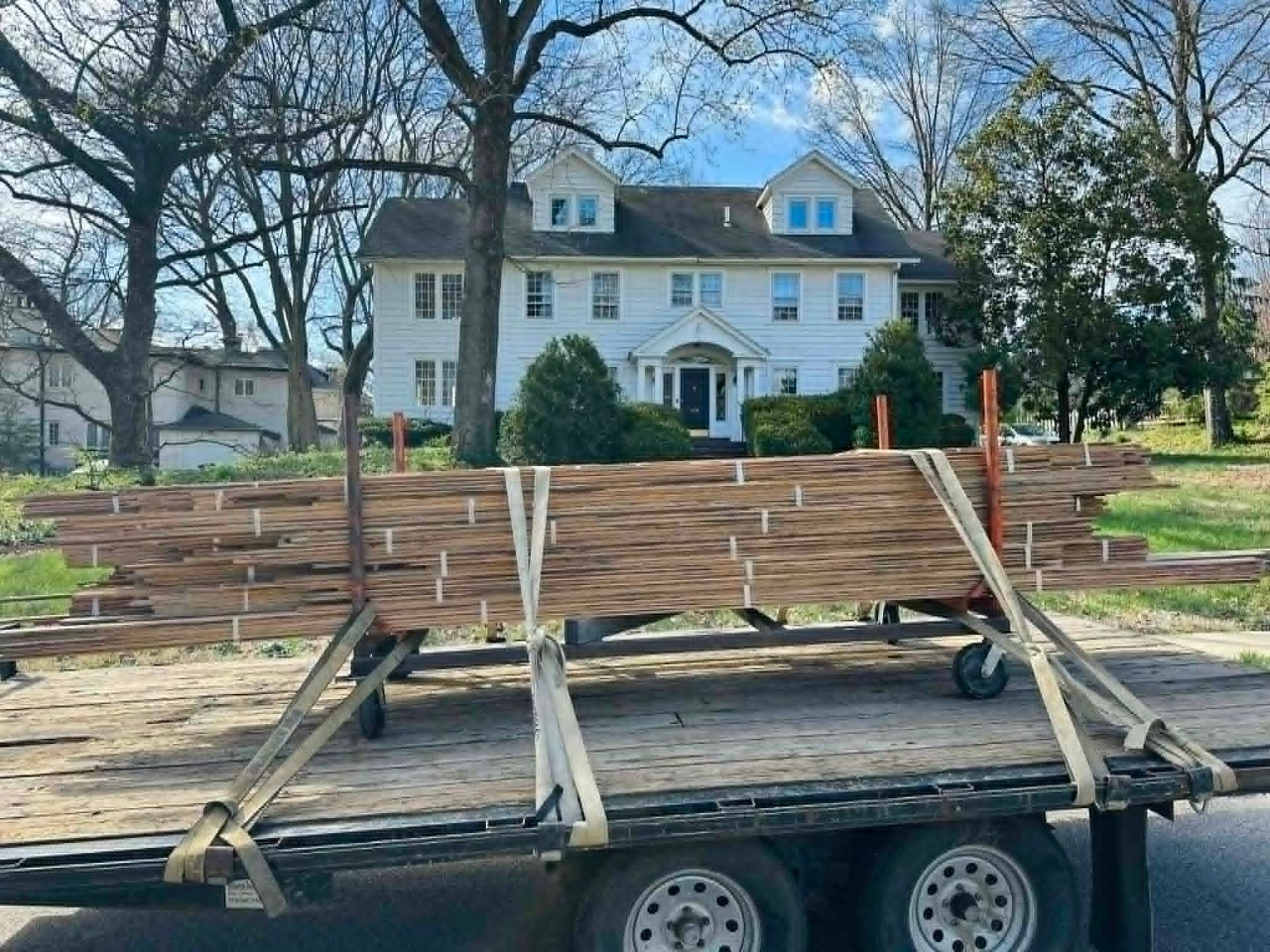 Flatbed trailer loaded with stacked wooden planks in front of a white house and bare trees