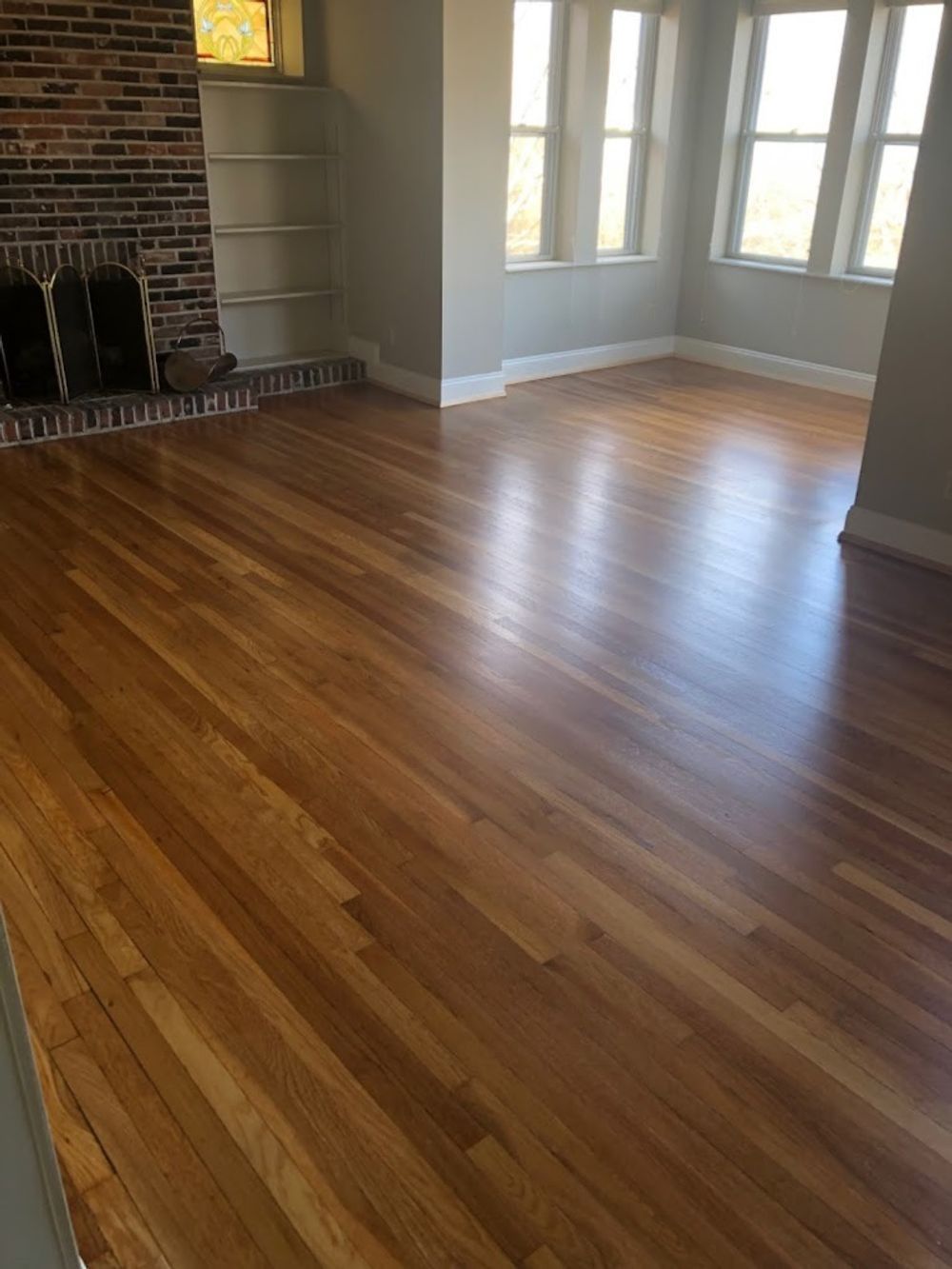 An empty room with polished hardwood floors, light gray walls, white trim, and a brick fireplace with built-in shelves.