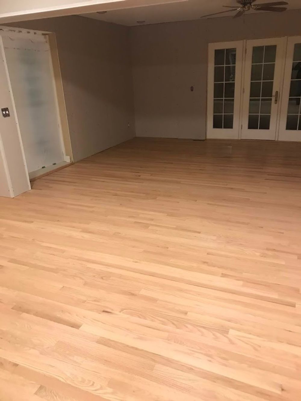 A wide, empty room featuring newly installed light-toned hardwood floors, grey walls, and white French doors.