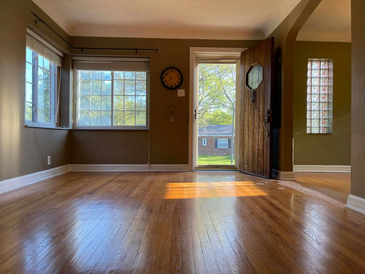 Empty living room with hardwood floors, large windows, and warm sunlight streaming through the doorway.