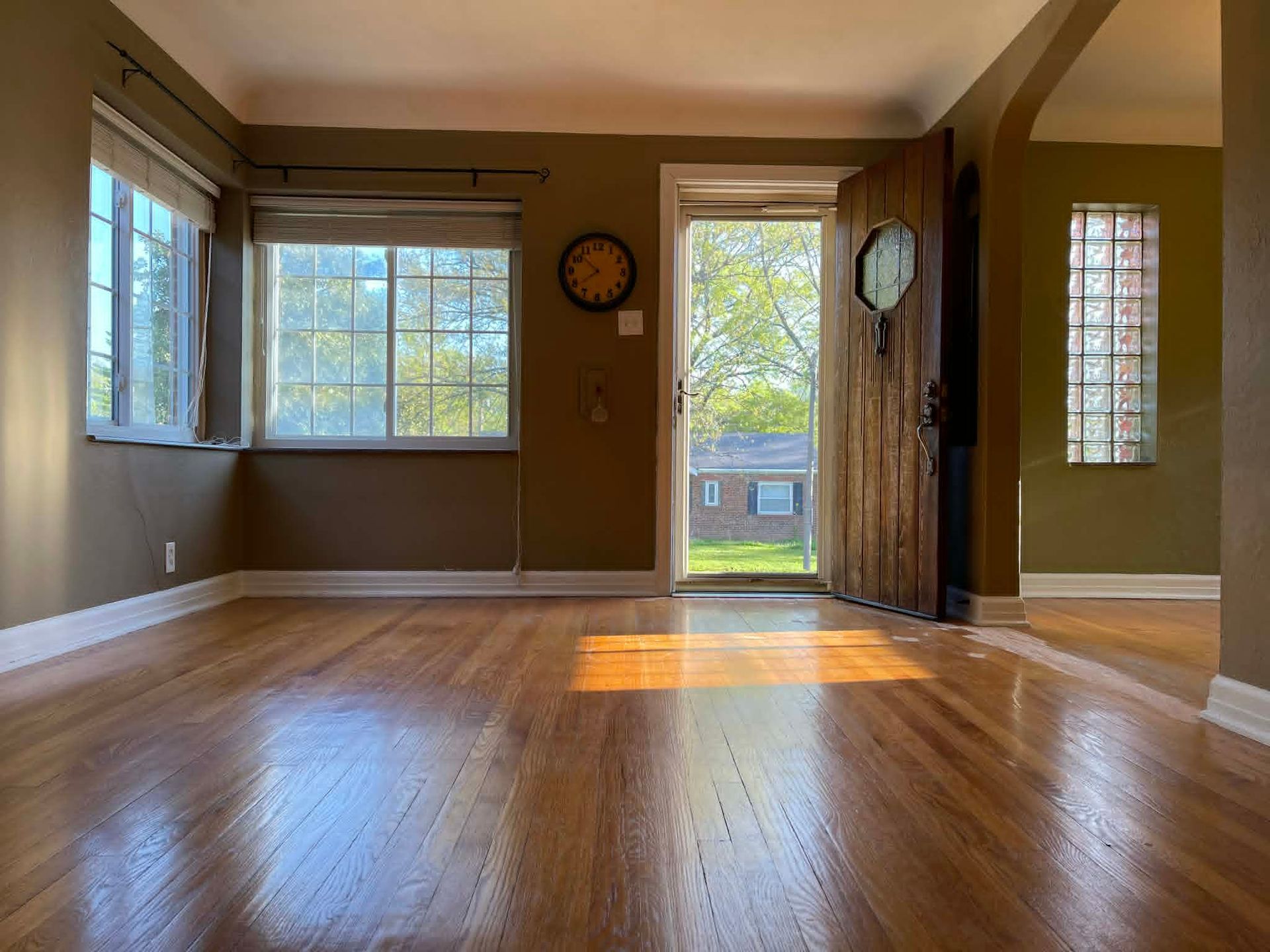 Empty living room with hardwood floors, large windows, and warm sunlight streaming through the doorway.
