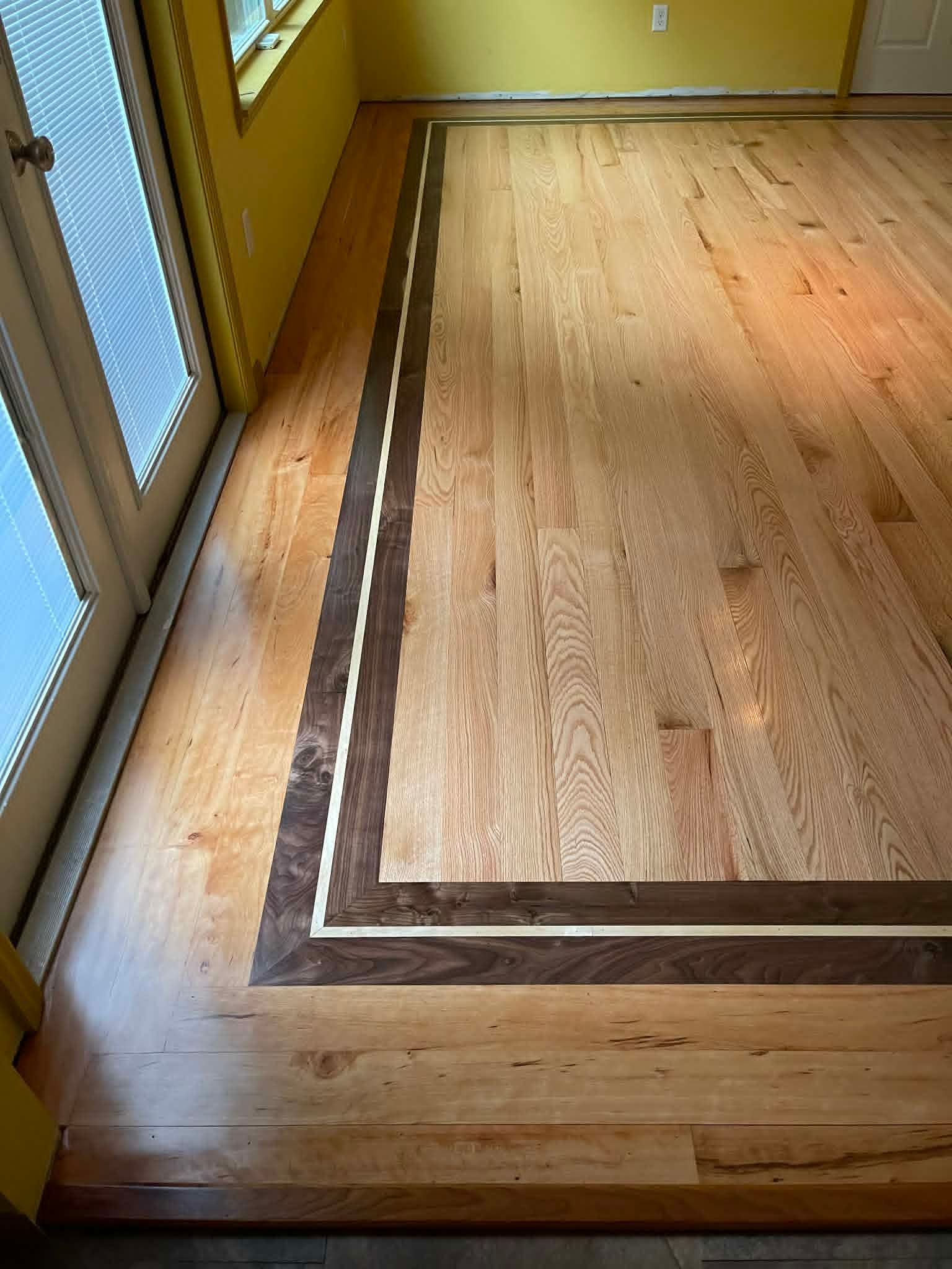 Wooden floor with dark border in a sunlit room beside a window and glass door.