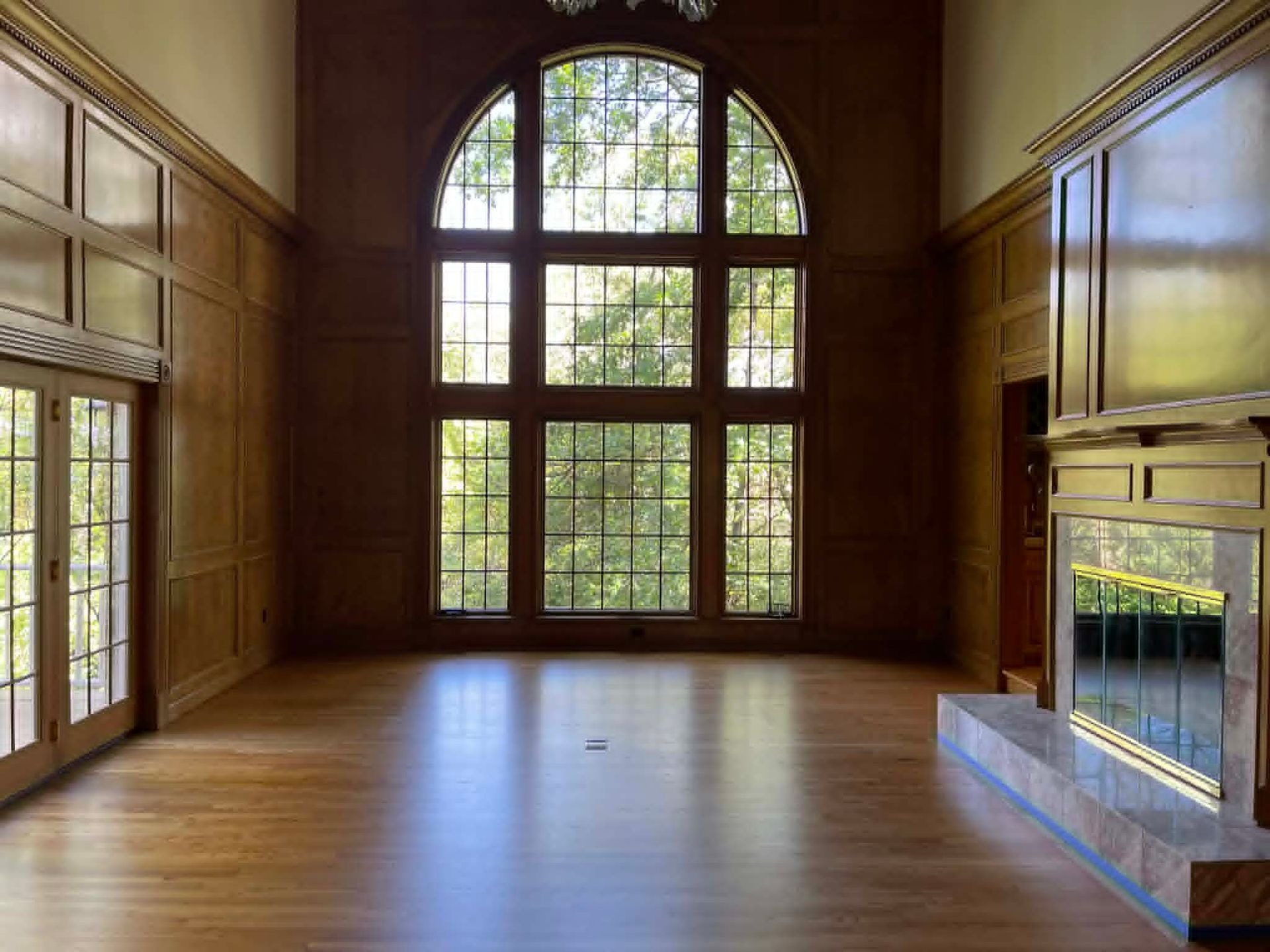 Empty wood-paneled room with large arched window and fireplace, filled with natural light