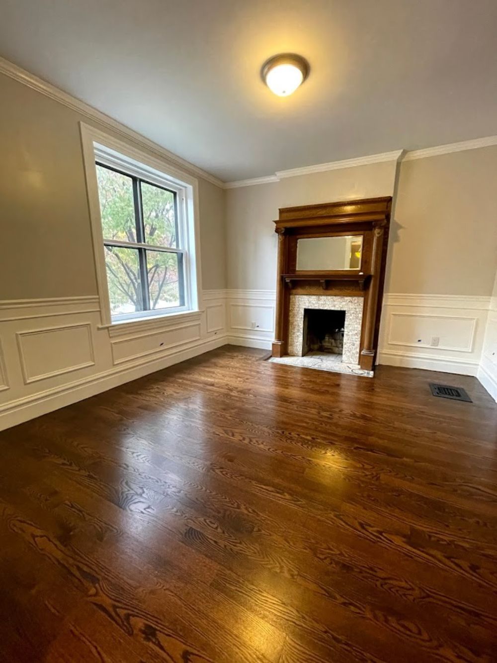 An empty room with dark hardwood floors, white wainscoting on gray walls, a large window, and a wooden fireplace mantle.