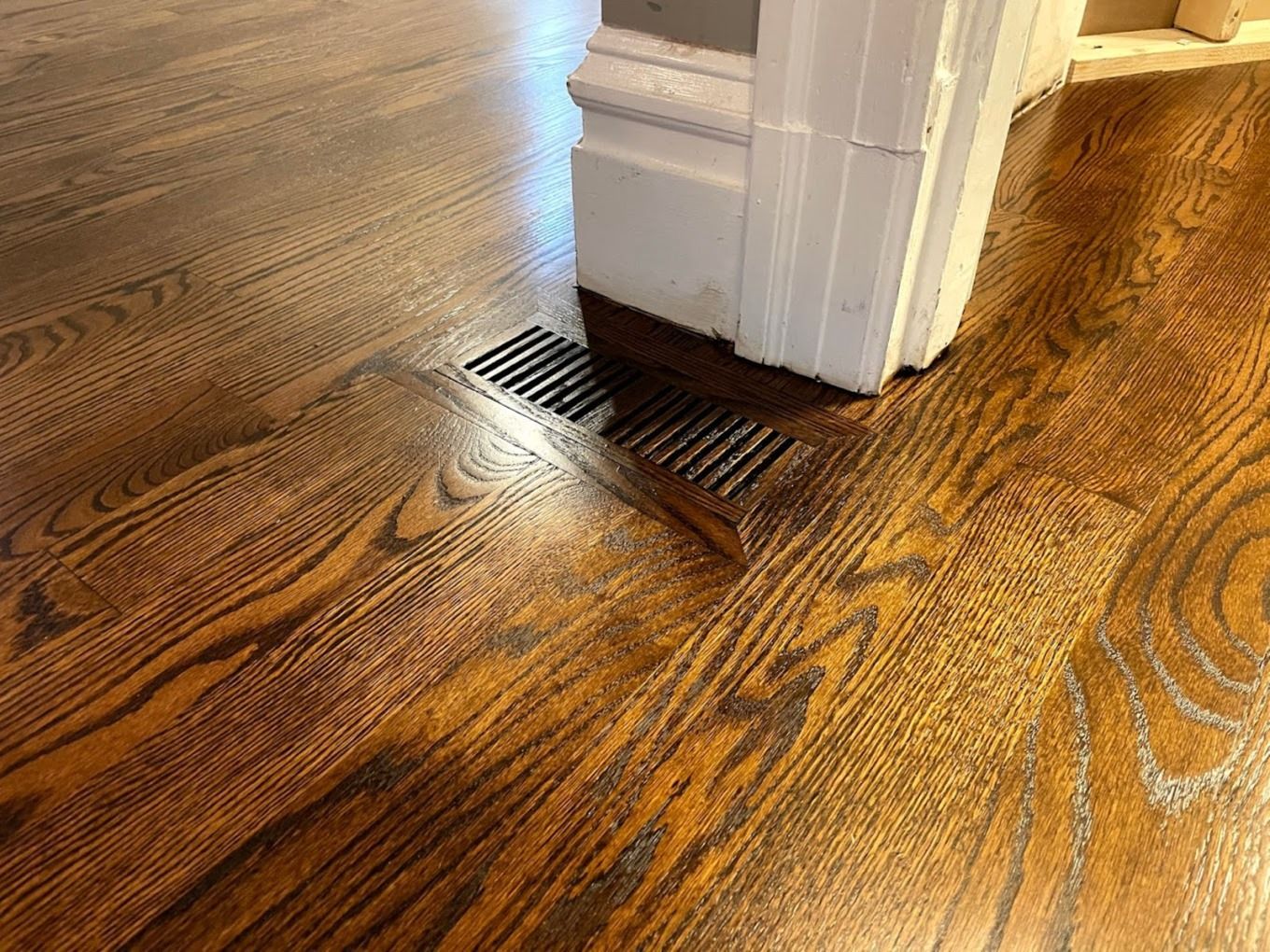 A black metal floor vent set into dark stained hardwood flooring, positioned against a white wooden door frame.