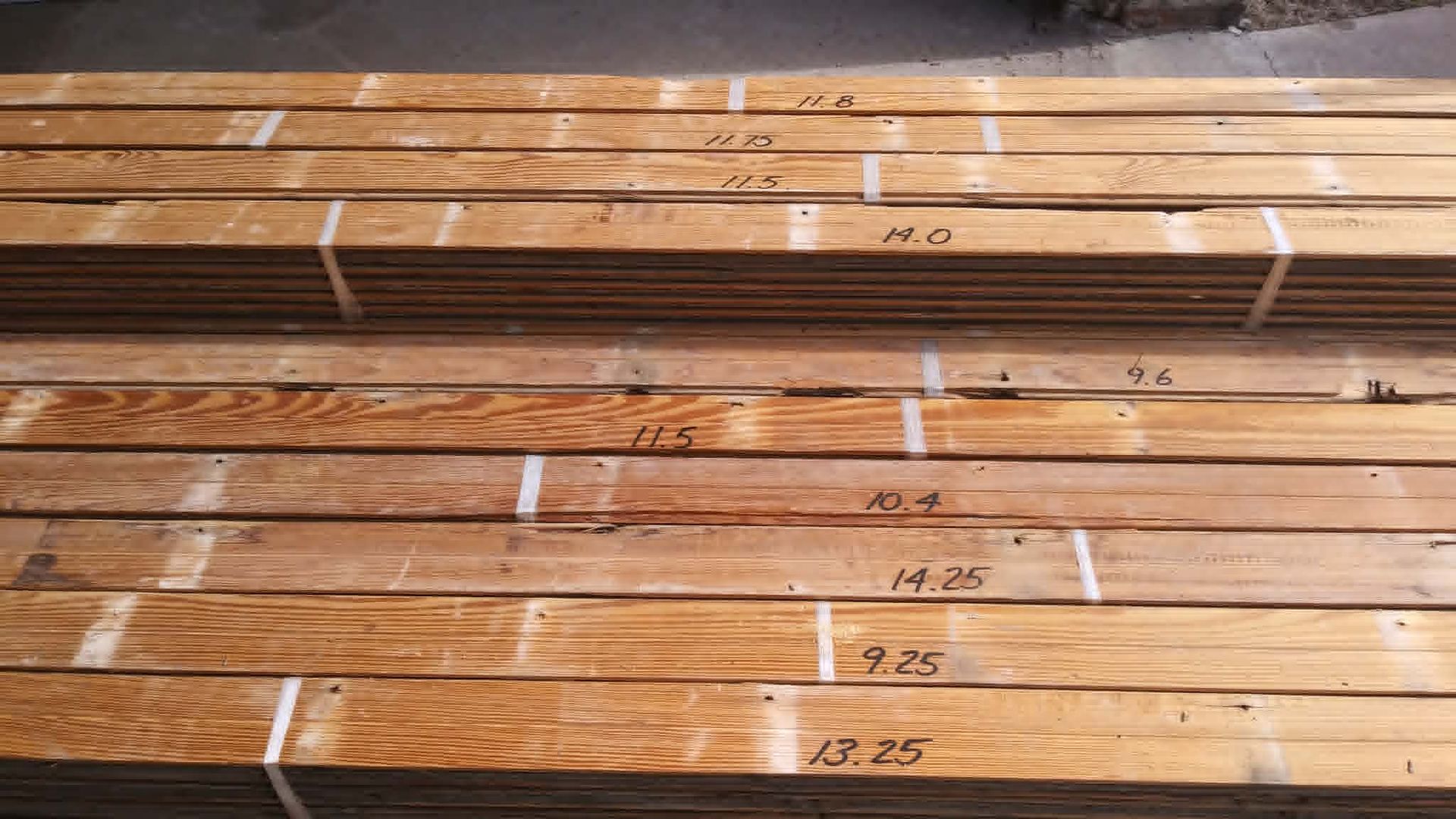 Stacked wooden planks with white markings, shown in a workshop or lumber yard.