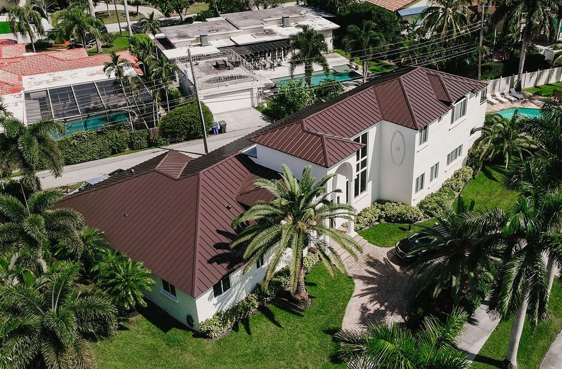 An aerial view of a large white house with a brown roof surrounded by palm trees.