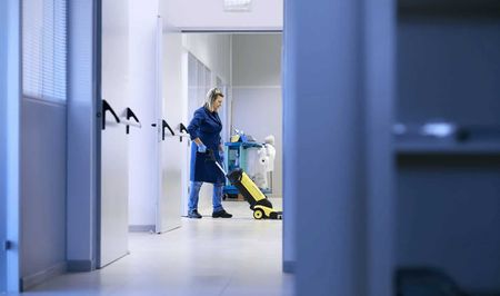 Person cleaning a hallway with a vacuum cleaner. A cleaning cart is nearby.