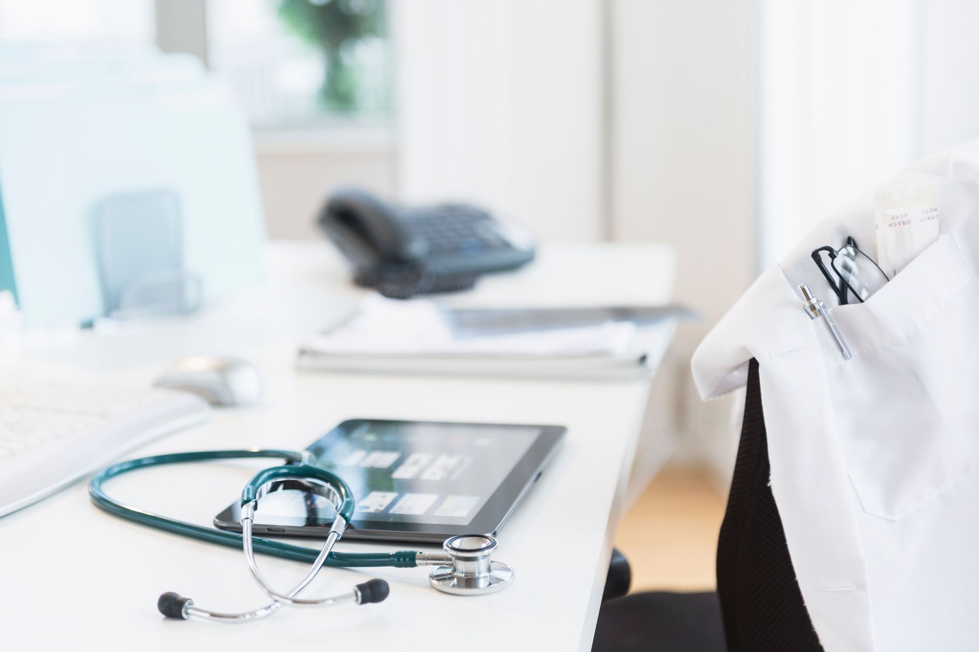 Stethoscope and tablet on a doctor's desk next to a white coat.