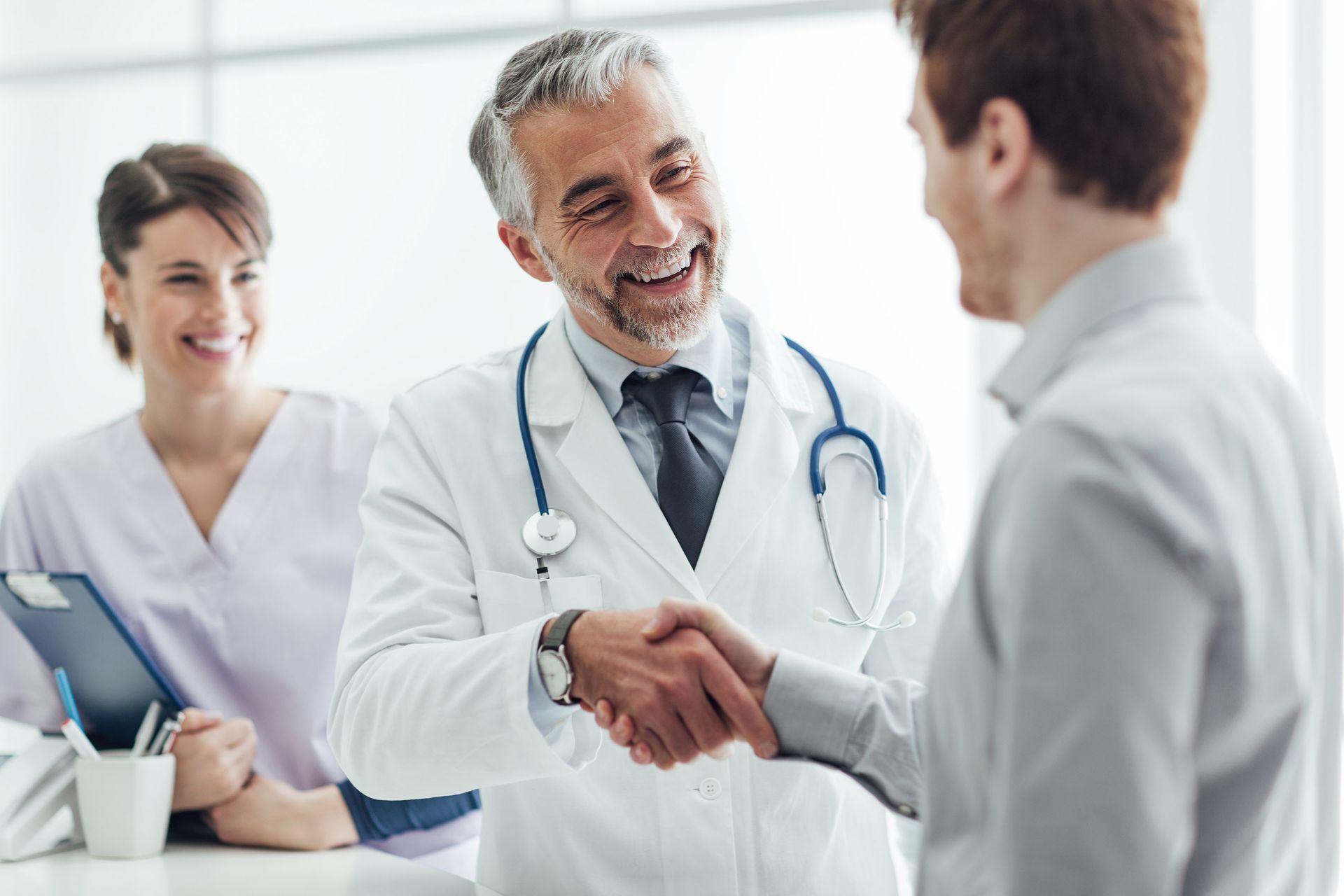 Doctor shaking hands with a patient, nurse smiling nearby. White coats, stethoscope, bright office setting.