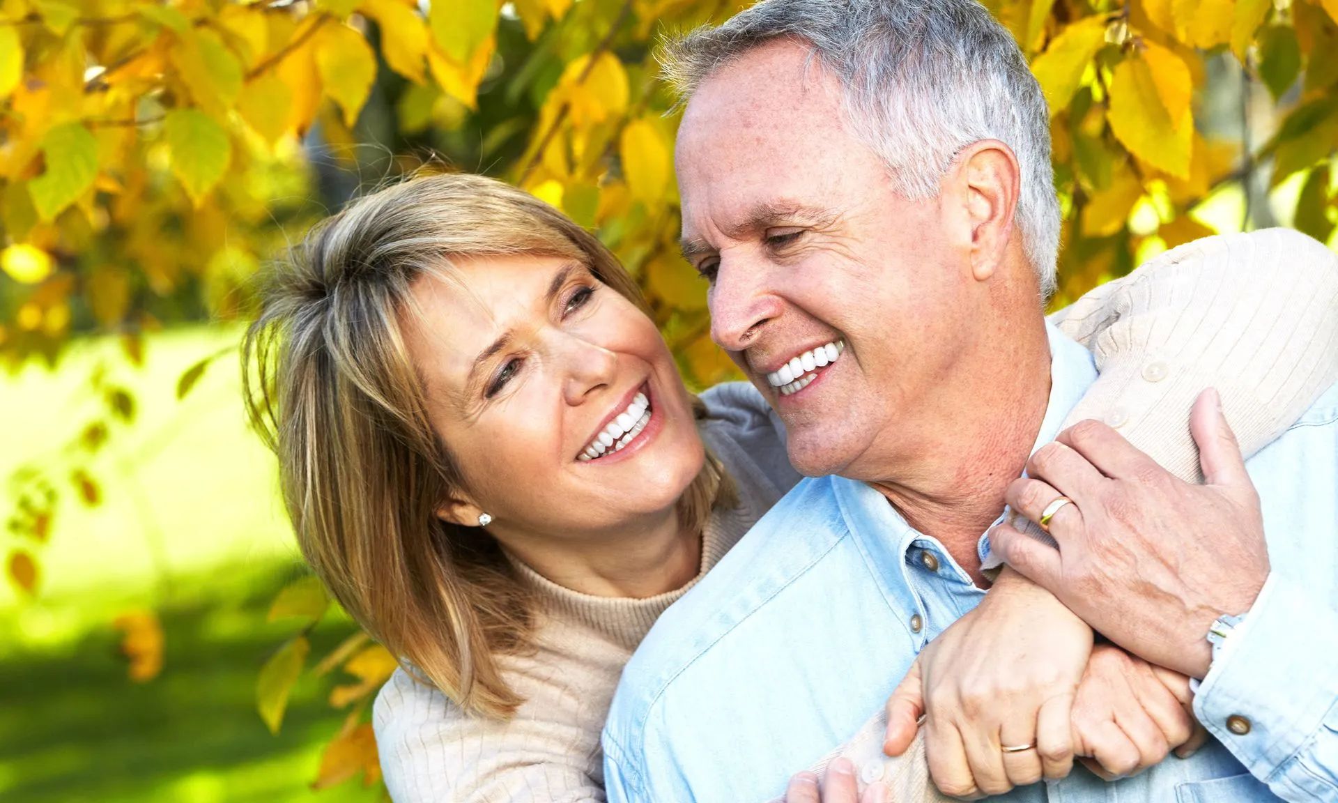 Couple smiling, embracing outdoors with yellow autumn leaves.