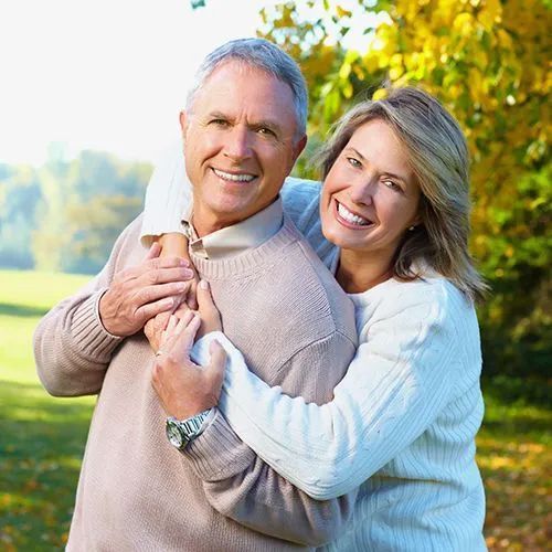 Woman hugging man outdoors, both smiling. Fall foliage in background.