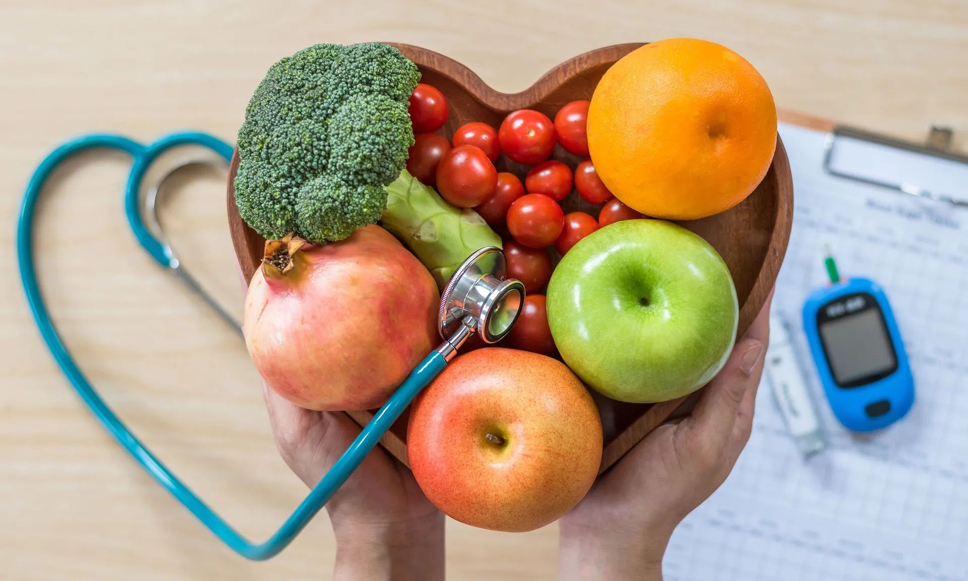 Hands holding a heart-shaped bowl filled with fruits and vegetables, stethoscope, blood glucose meter, and clipboard.