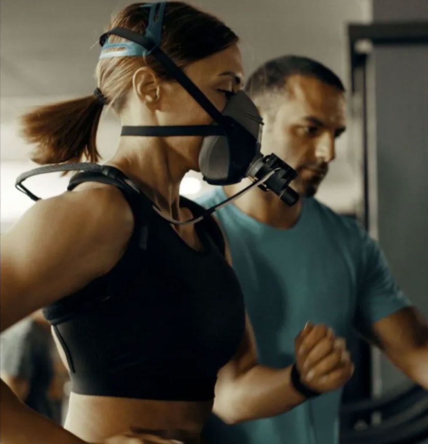 Woman running on treadmill with mask and equipment, being monitored by a trainer in a gym setting.