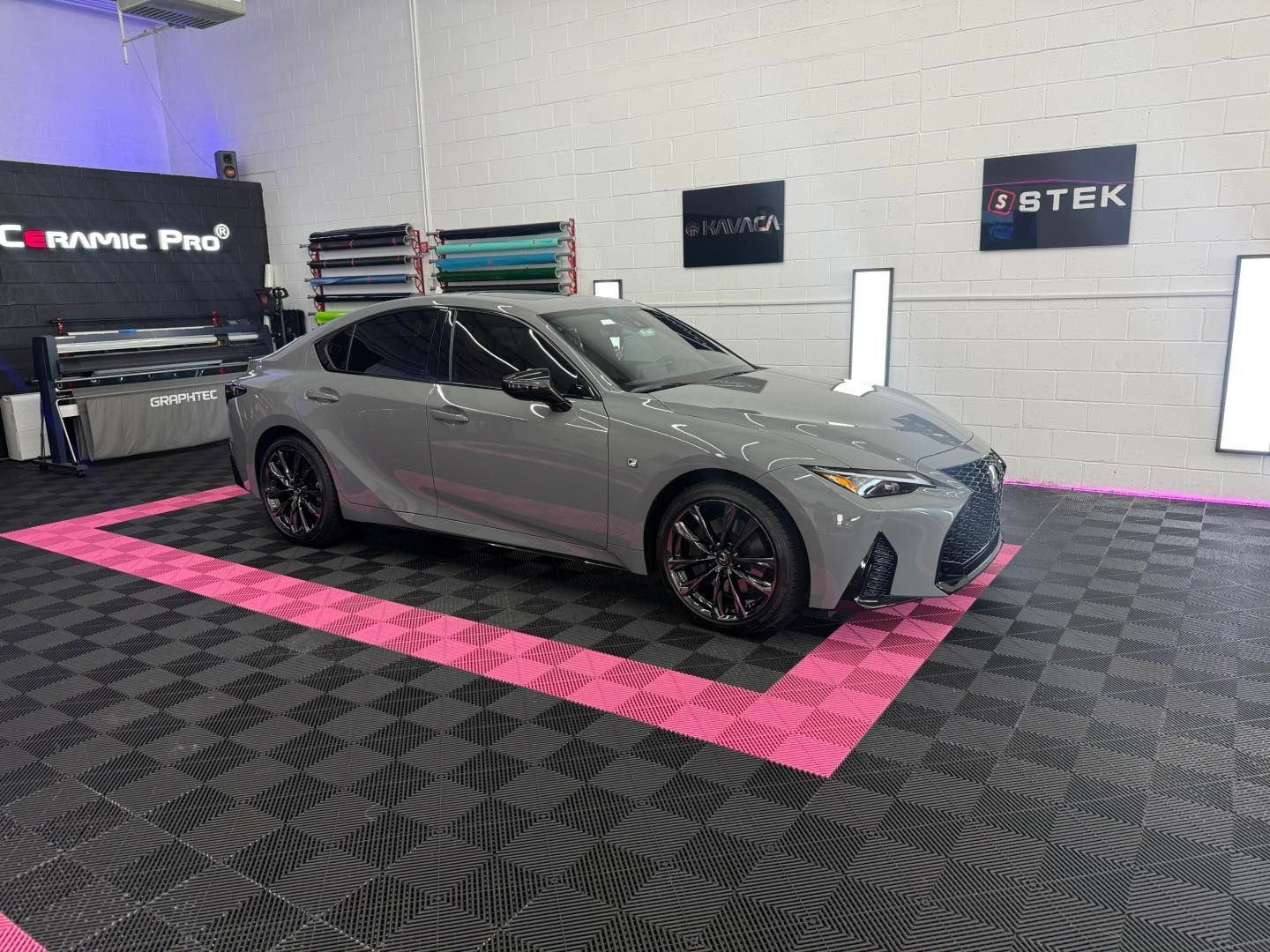 Gray Lexus sedan inside a car wrapping shop on a checkered floor with pink borders.