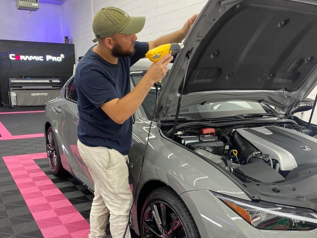 Man using a heat gun on a car hood in a shop. The car is gray, and the man wears a cap and khaki pants.