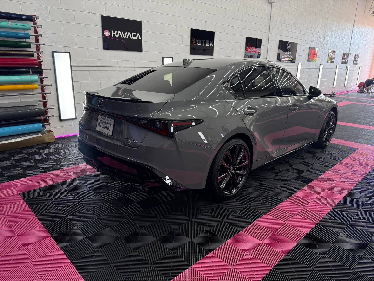 Gray Lexus sedan parked on a black and pink tiled floor, inside a shop. Rolls of colored vinyl are visible on the left.