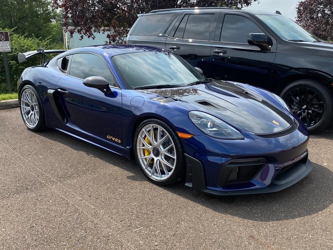 Blue Porsche 718 GT4 sports car parked outside. Black roof accents, silver wheels, yellow calipers.