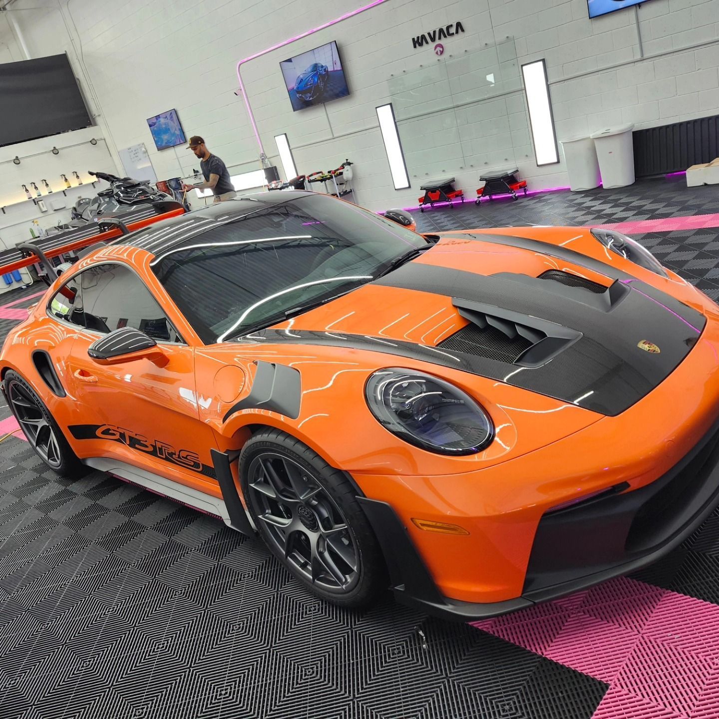 Bright orange Porsche 911 in a garage with black accents, black wheels, and a person in the background.