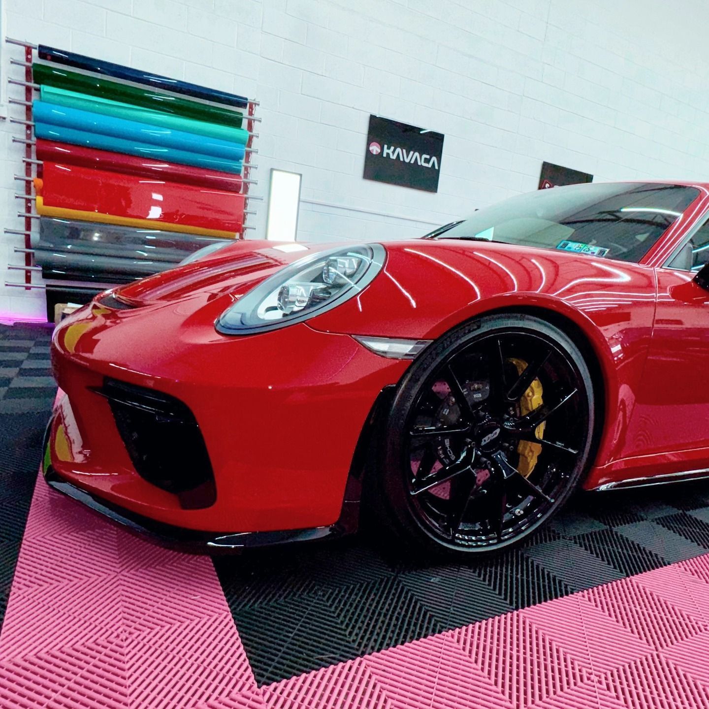 Red Porsche 911 with black wheels in a shop, rolls of colored vinyl on wall.