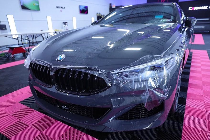 Dark gray BMW with clear protective film on front, in a garage with pink and black floor tiles.