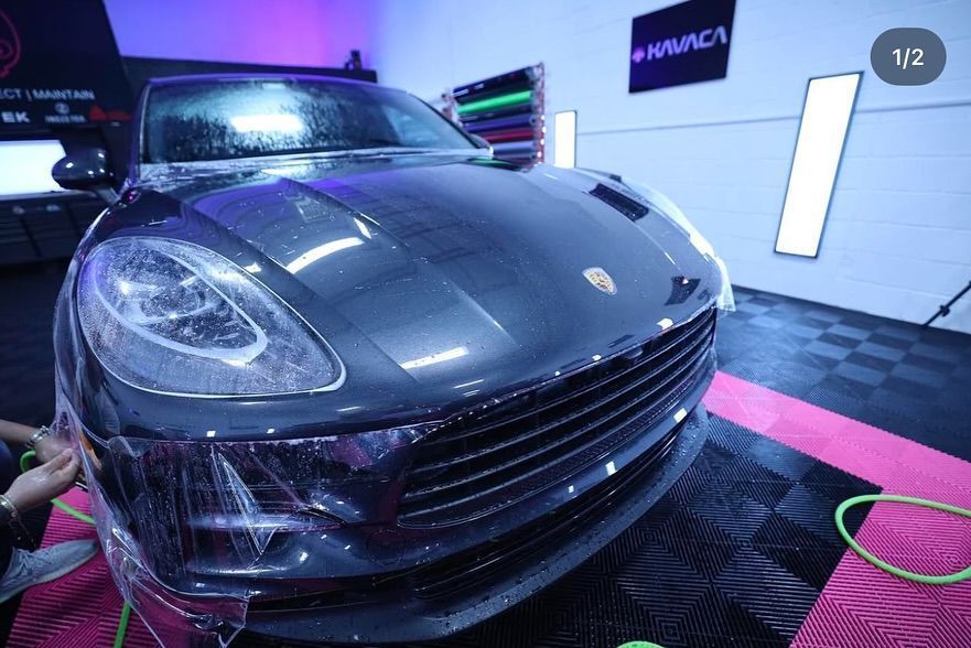 A person applying film to a dark gray Porsche Macan in a brightly lit garage.