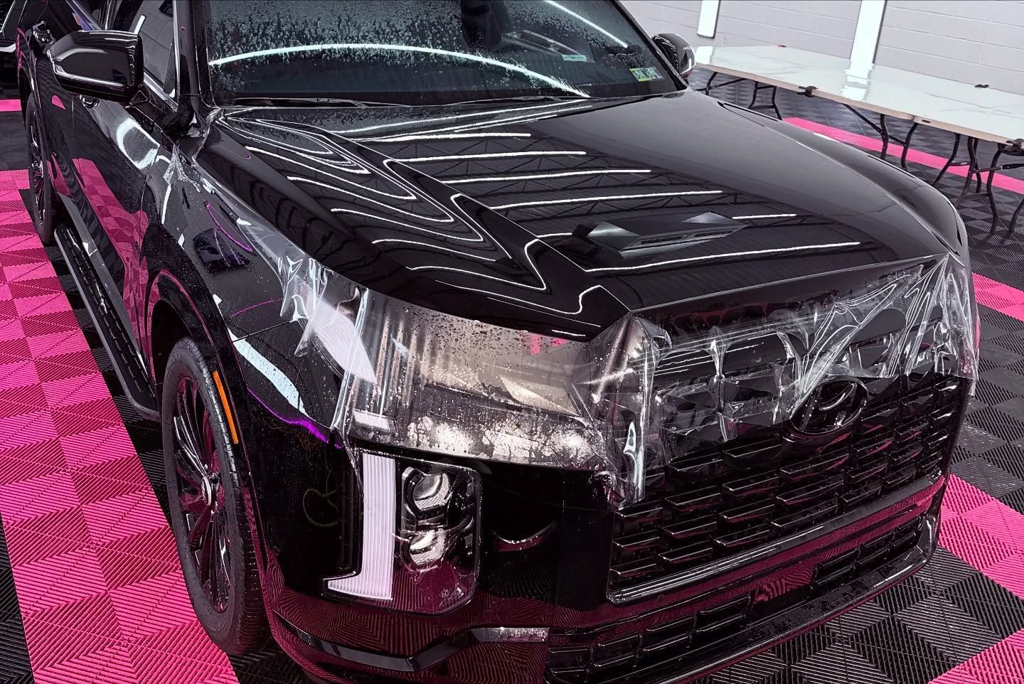 Black SUV, front view. Clear protective film on hood and grill, glossy finish, parked on pink and black tile.