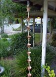 A copper rain chain is hanging from a porch in a garden.