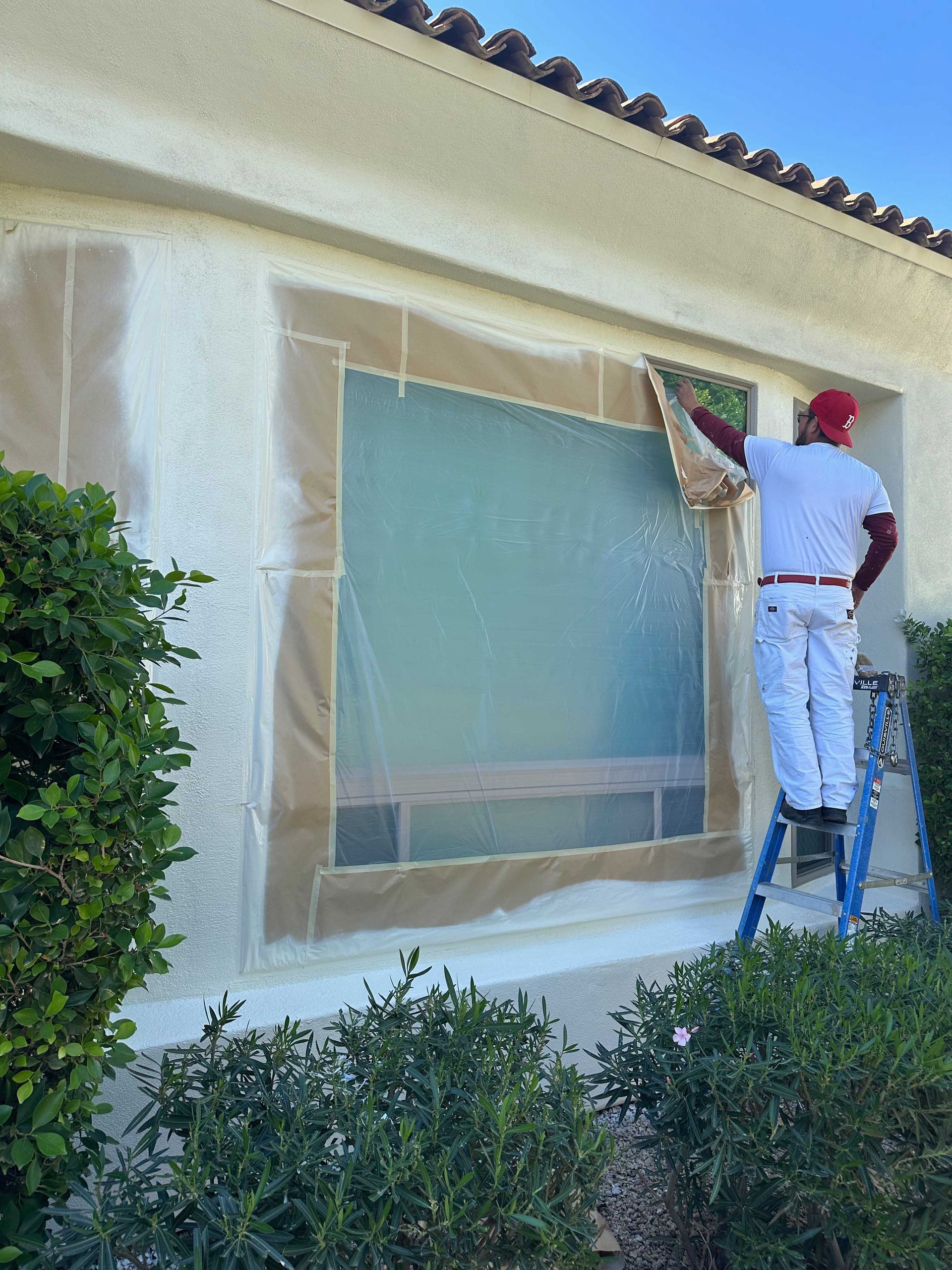 A man is standing on a ladder painting a window of a house.