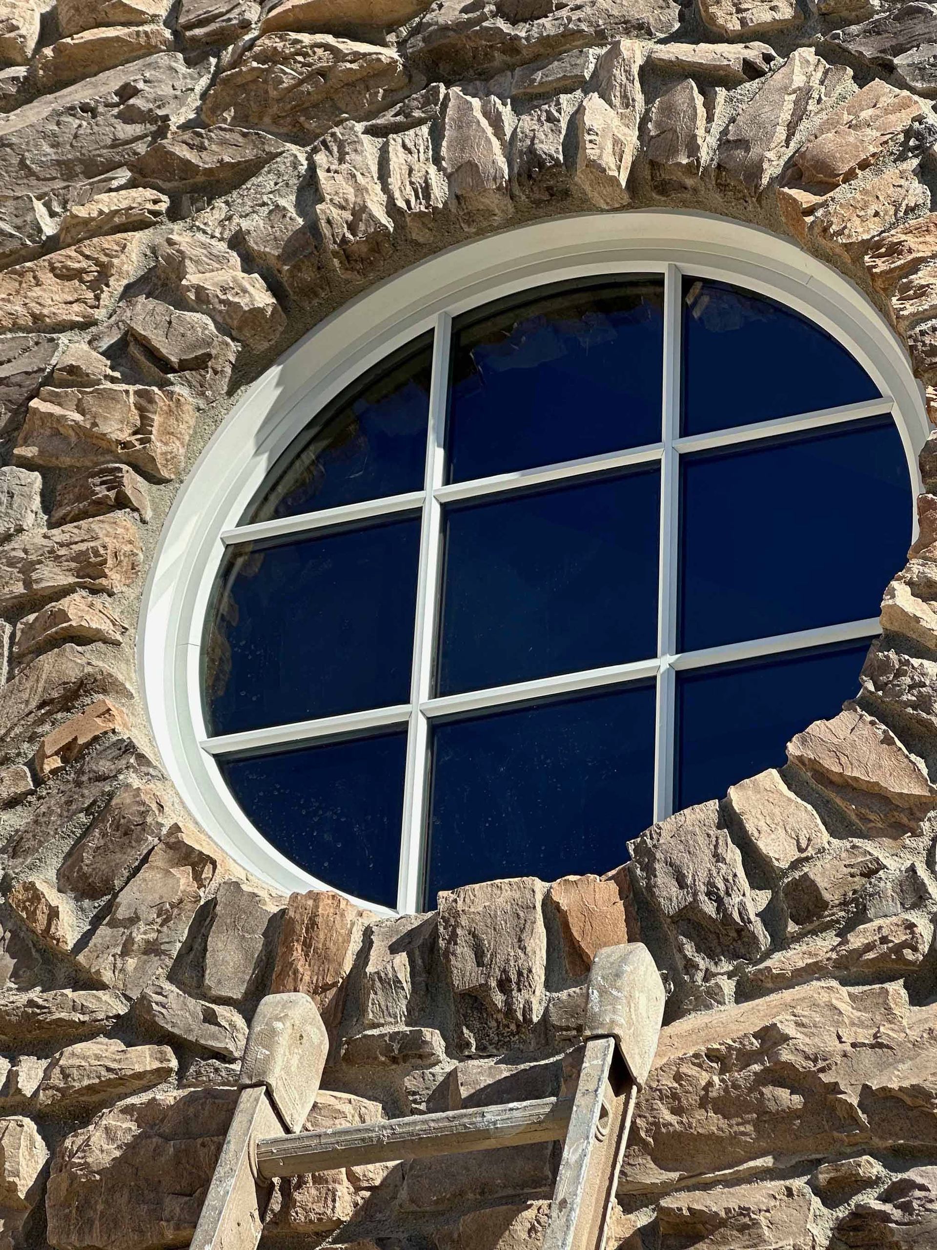A round window in a stone wall with a ladder in front of it.