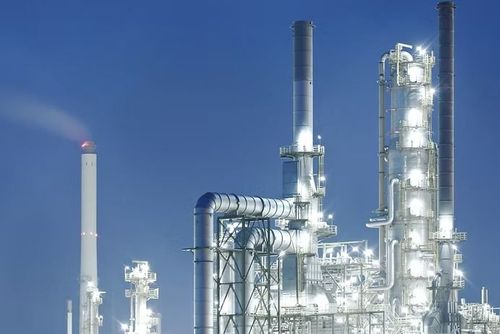 Oil refinery at night, illuminated, with smokestacks and pipes against a blue sky.