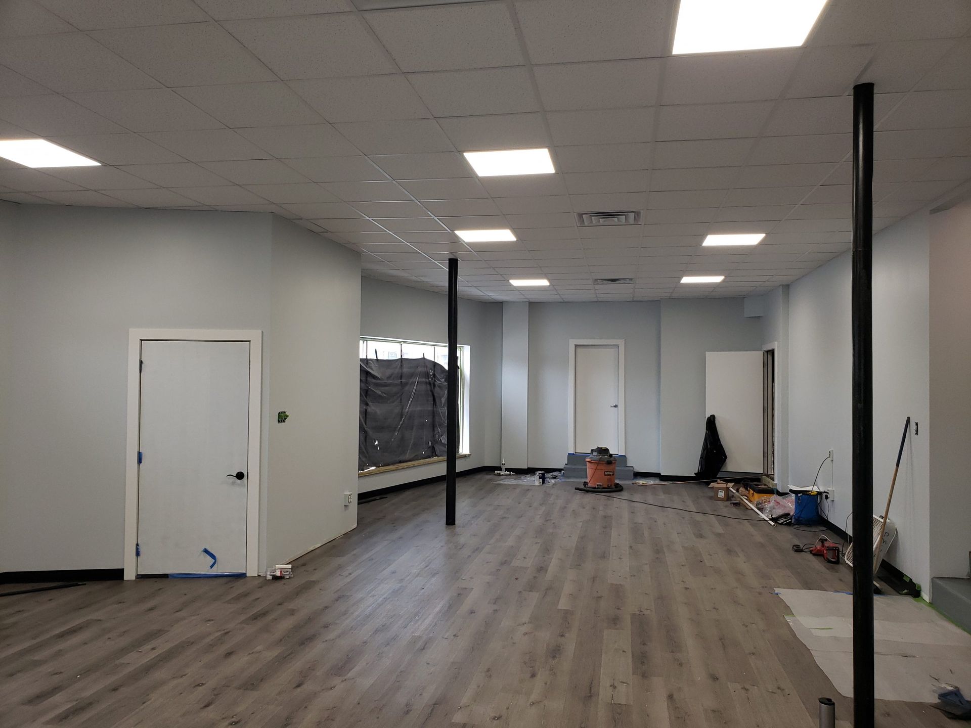 Interior of a mostly empty room with gray wood-look flooring and light-colored walls. Two black support beams are visible.