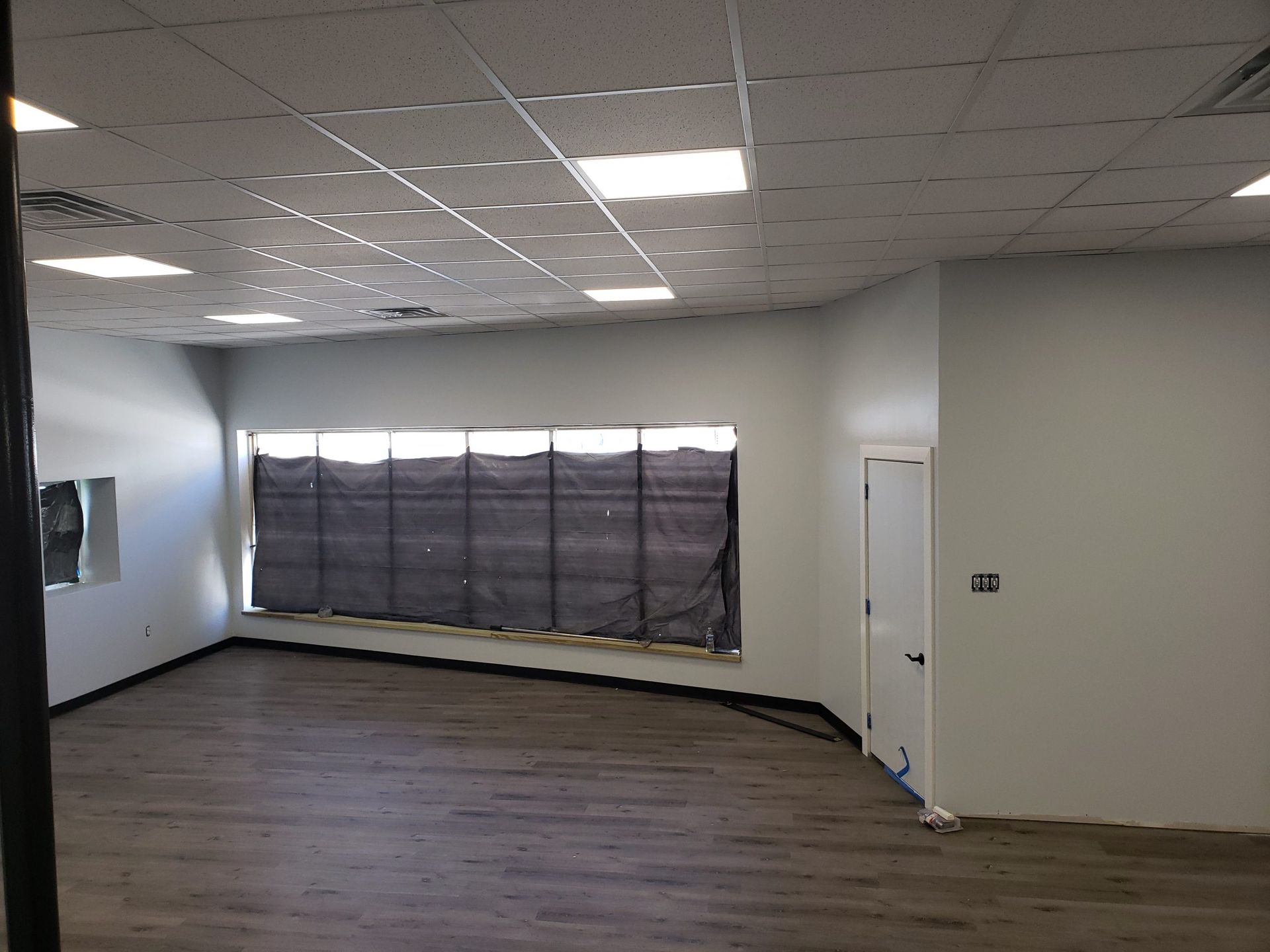 Empty interior of a room with gray walls, a window covered by a black shade, and wooden floors.