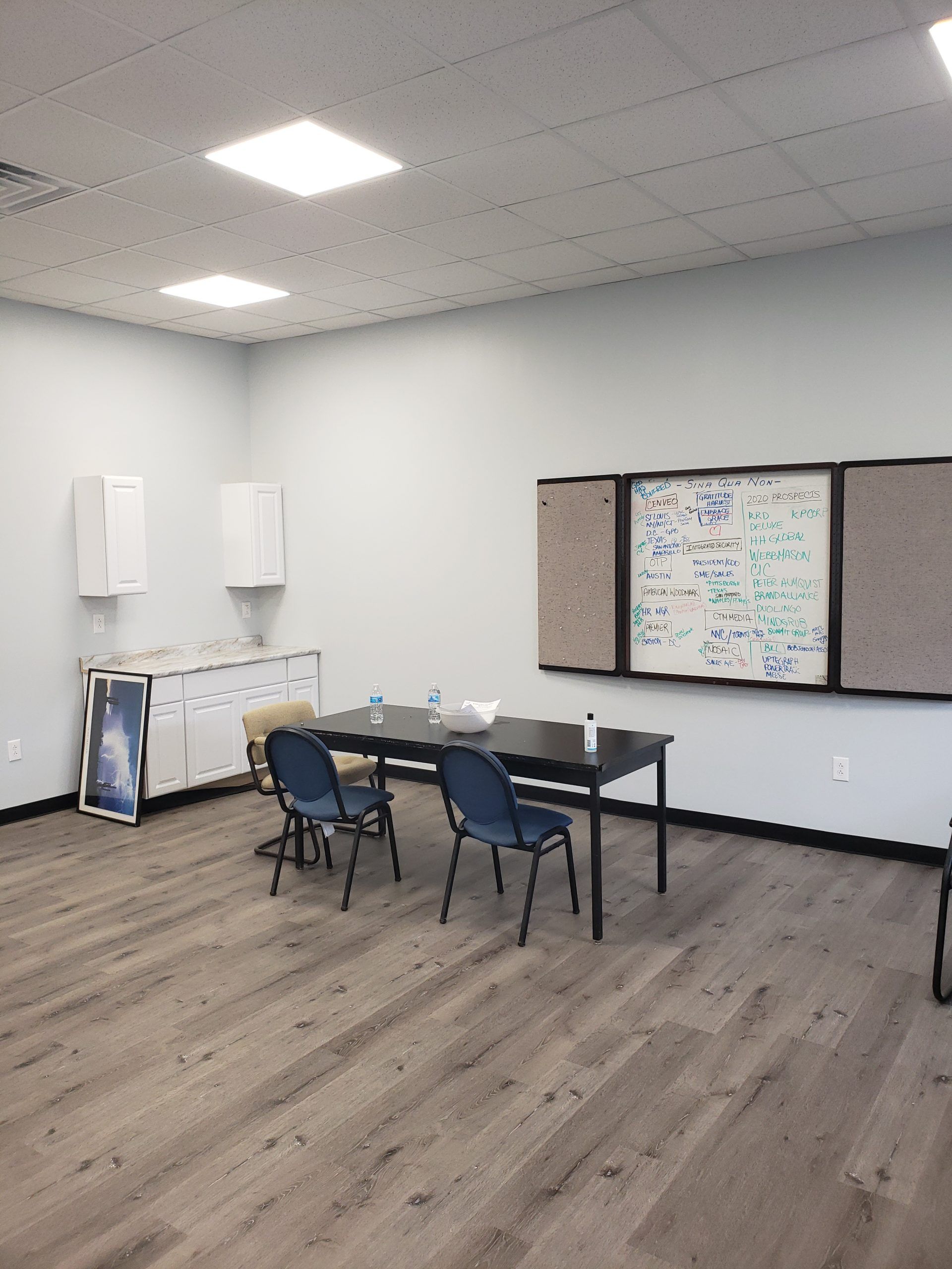 Empty room with table and chairs. Bulletin board, white storage cabinet, and laminate flooring.