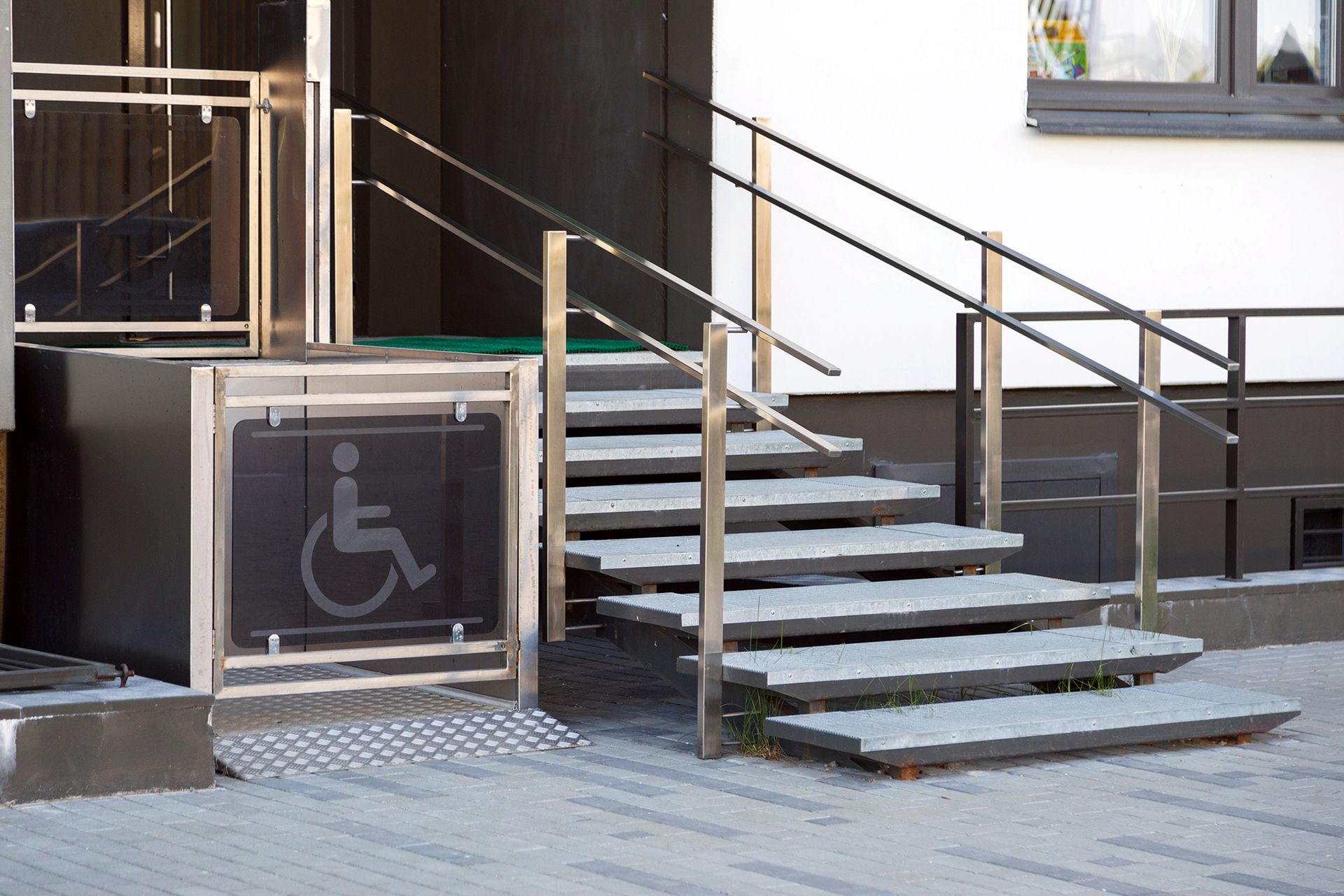 Wheelchair lift and stairs at building entrance. The lift has a disability symbol. Gray brick pavement.