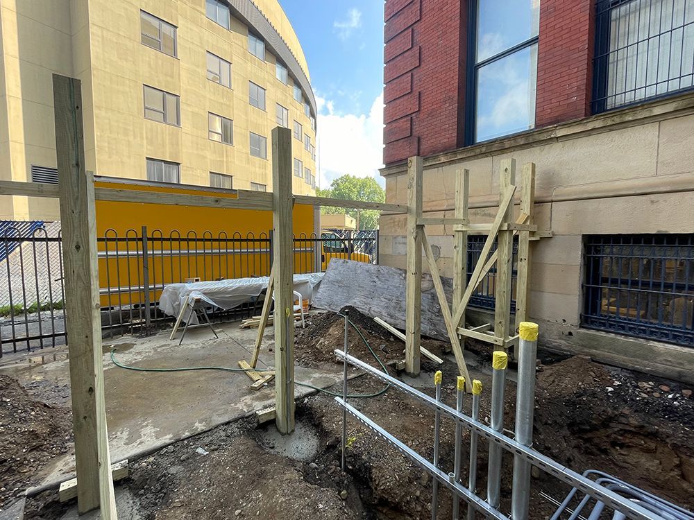 Construction site with wooden supports, next to a building and metal railing. Ground excavated.