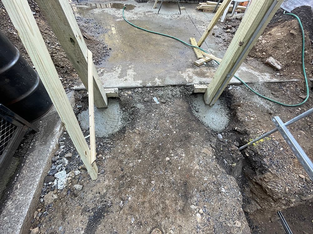 Wooden supports set in concrete, ground surrounding with a hose and metal fence visible.