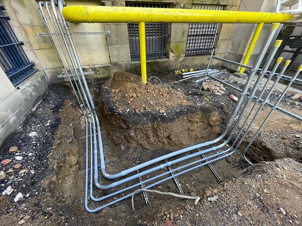 Construction site: exposed dirt, grey conduit pipes, yellow safety railing, exterior building wall.