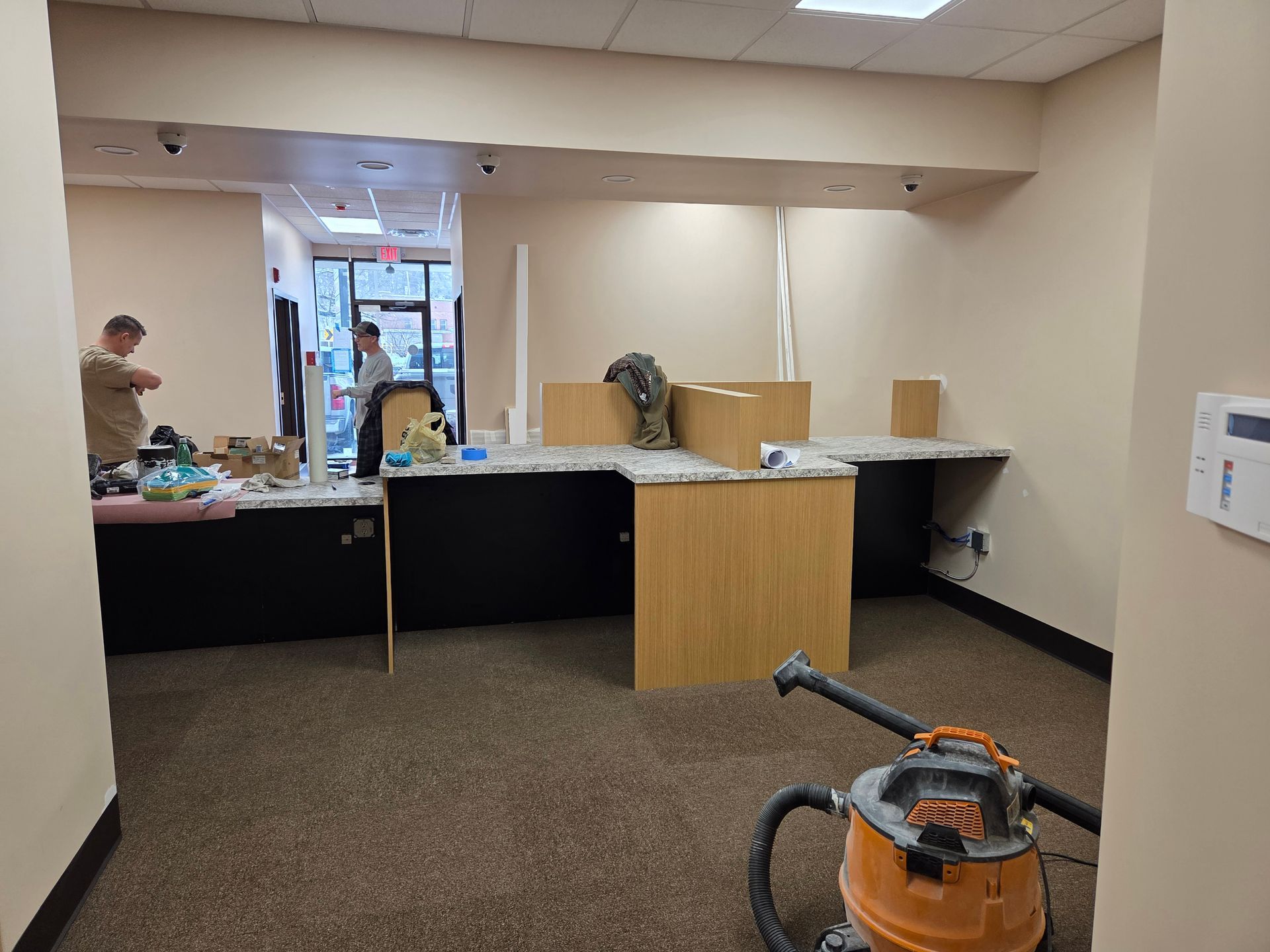 Interior of an office under renovation. Two people work at desks with beige walls, brown carpet, and a shop vac.