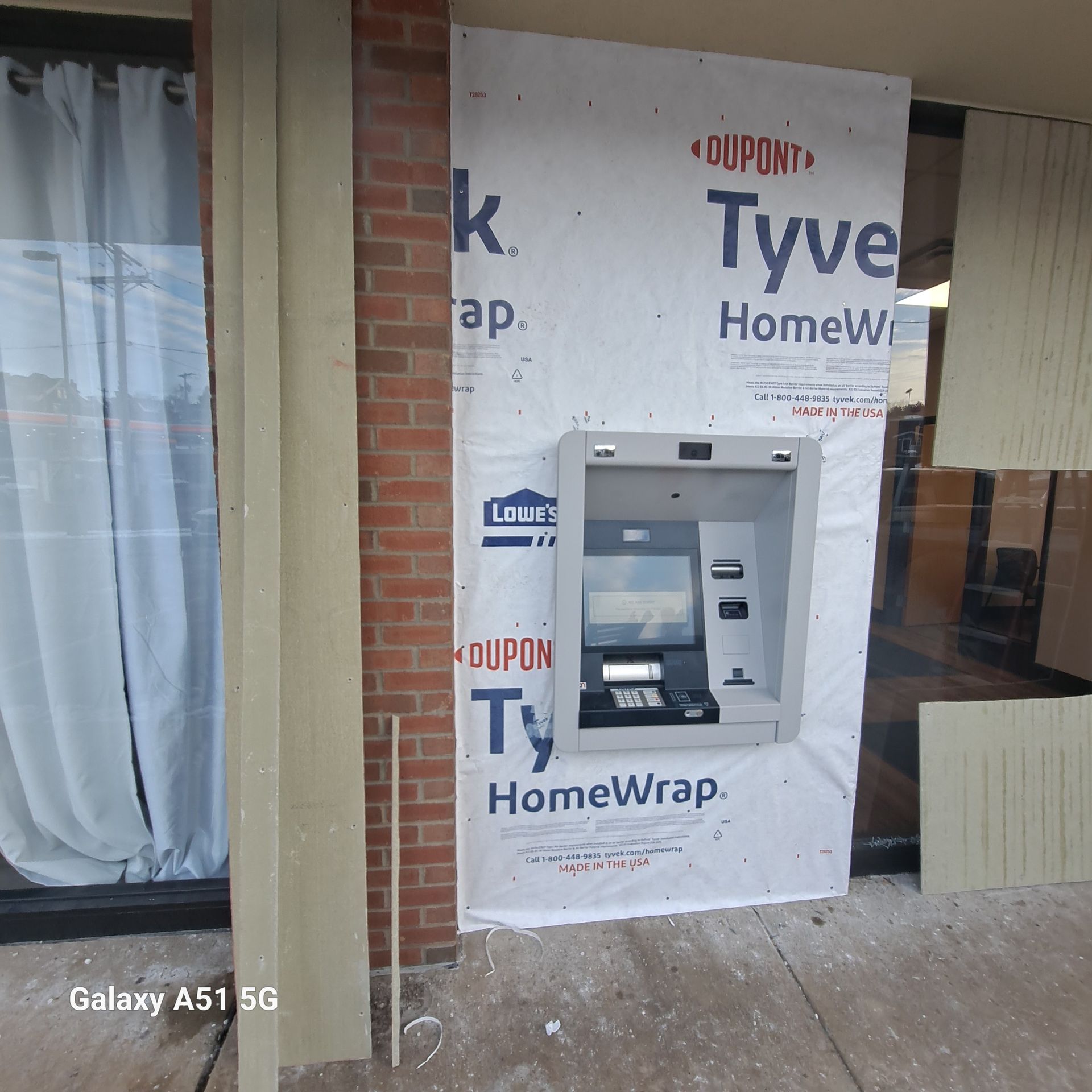 ATM installed on a building with Tyvek wrap. Bricks, curtains, and broken paneling are visible.