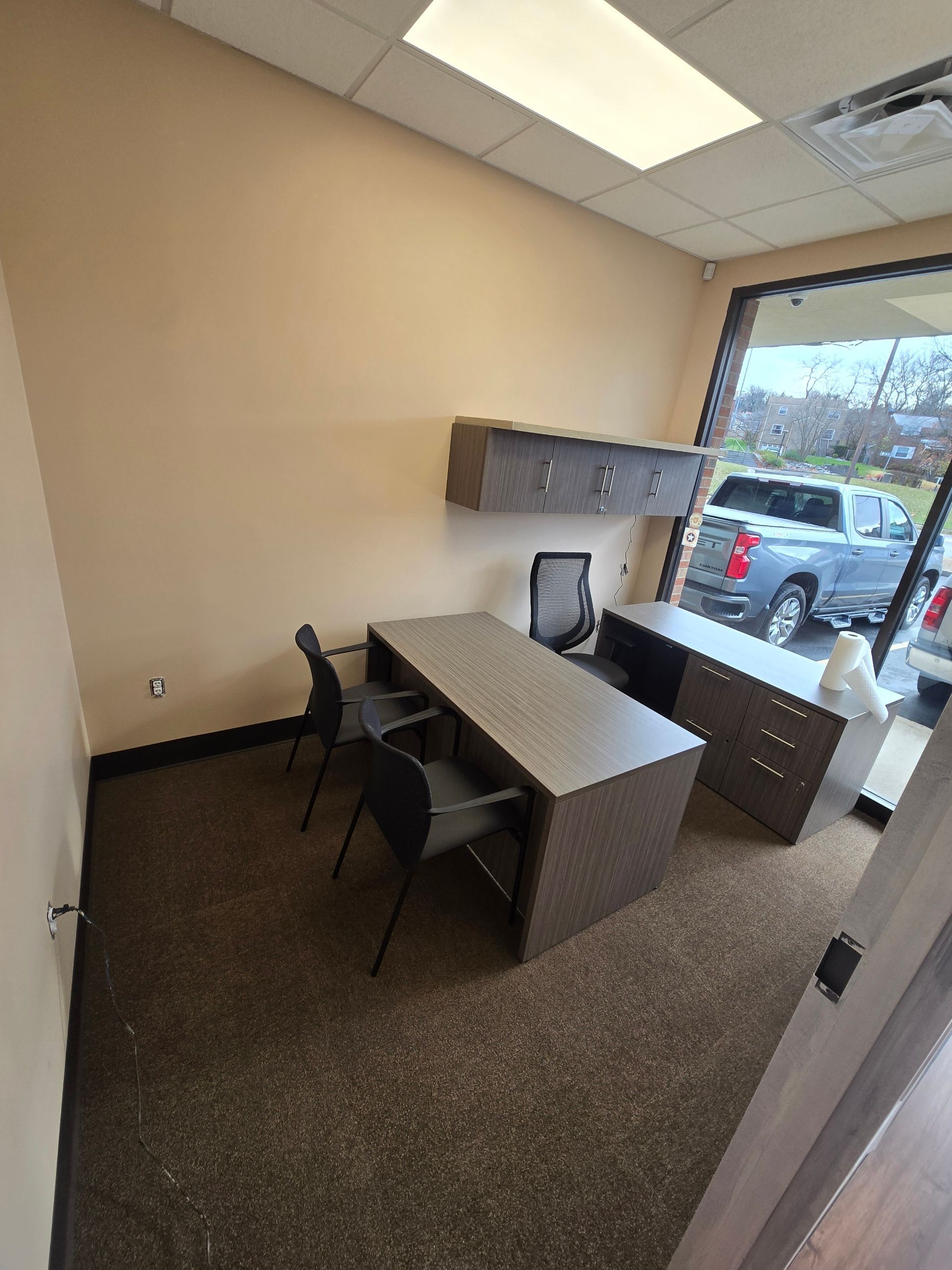 Office interior with desk, chairs, and cabinets. Window view of vehicles outside.