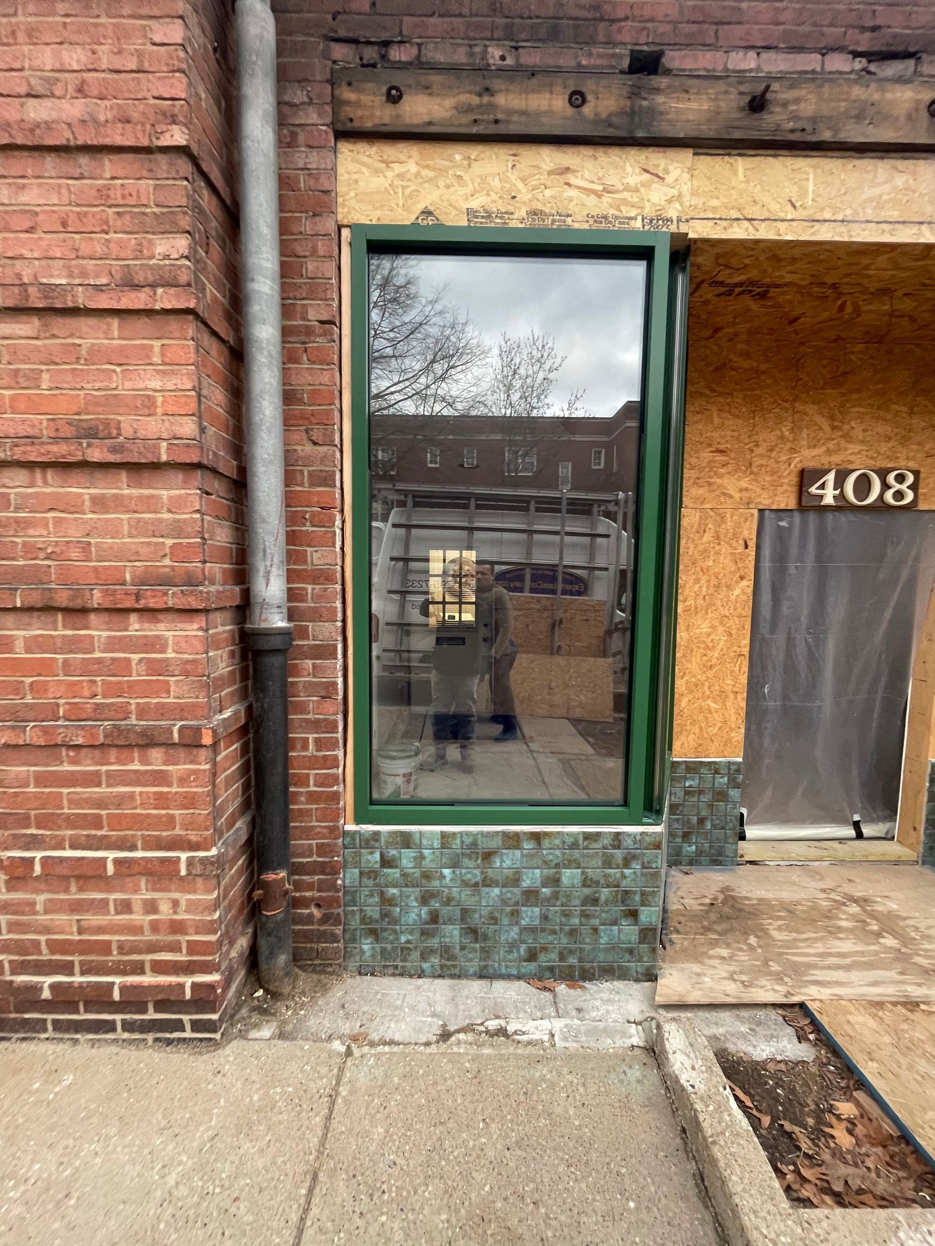 Storefront with green-framed window, brick wall, and mosaic tile base. Number 408 on door.