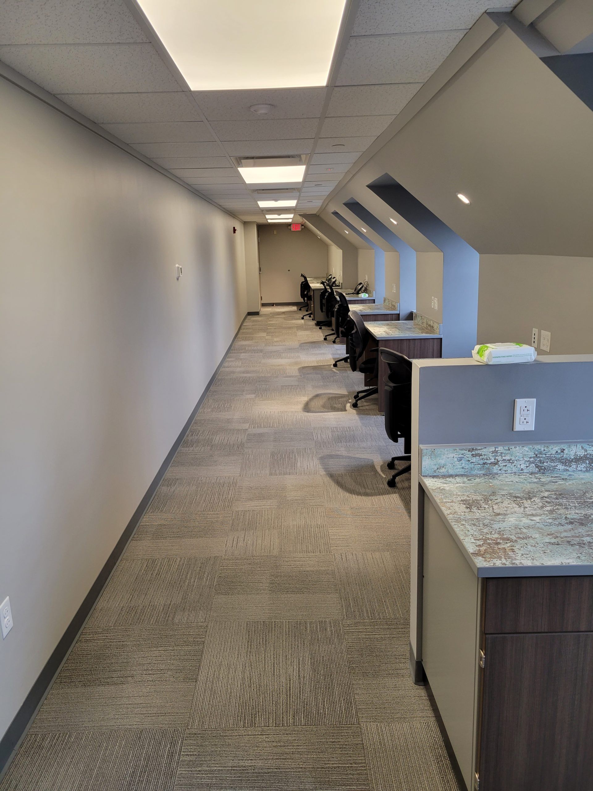 Long office hallway with desks, chairs, carpet, and neutral-colored walls. Fluorescent lights and an exit sign are visible.