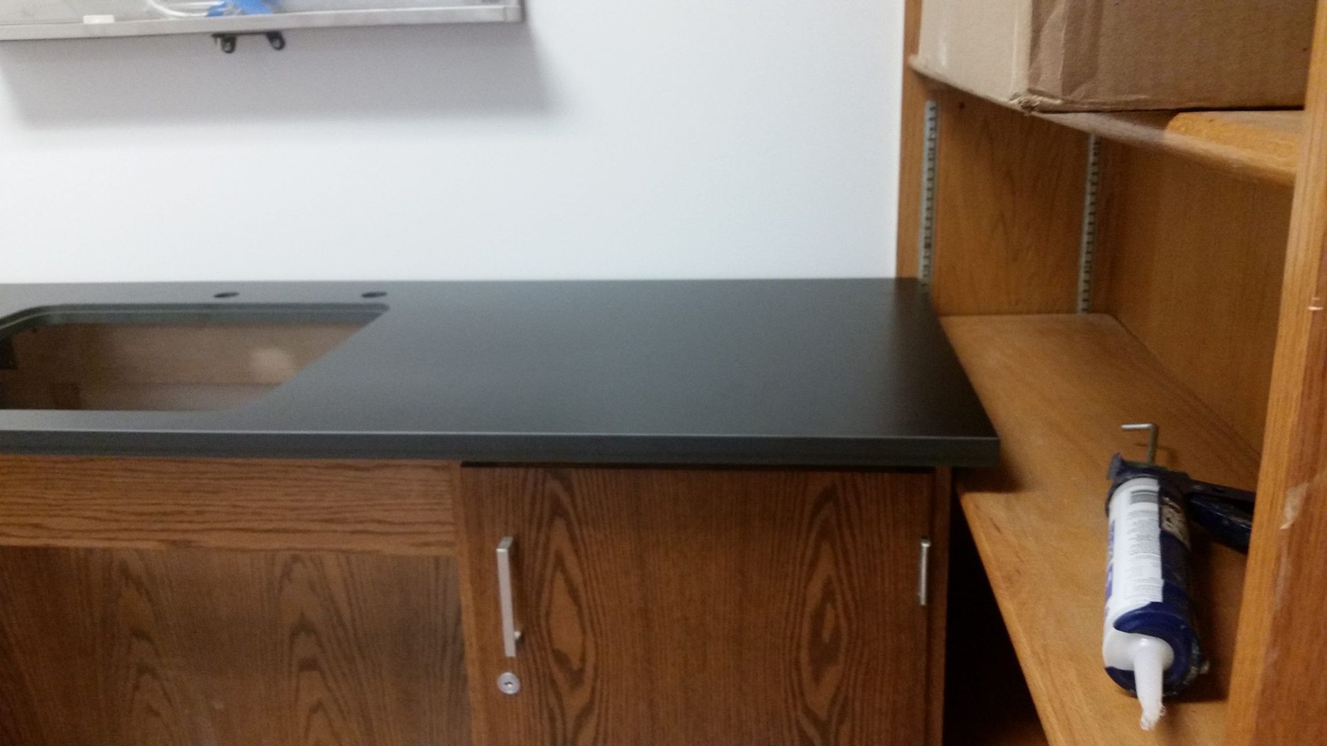 Countertop with sink, wooden cabinets, and shelving unit. Gray countertop, brown cabinets.