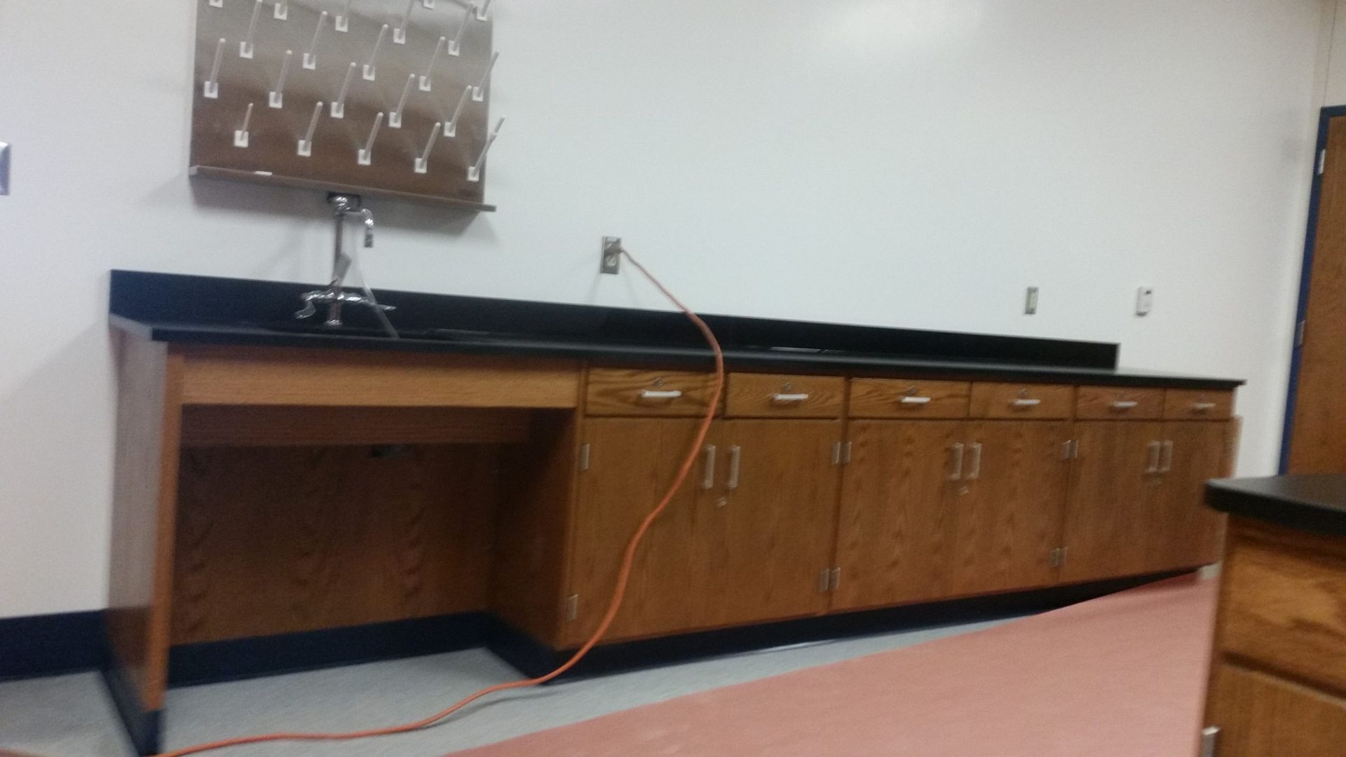 Wooden lab counter with a sink, drying rack, cabinets, and a power cord against a white wall.